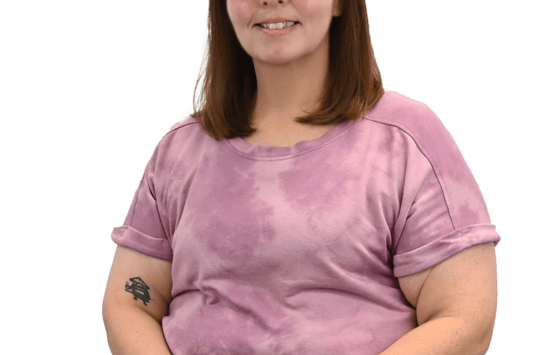 Alicia Robinson, a woman with brown hair and glasses wearing a pink t-shirt, smiles at the camera in a professional portrait