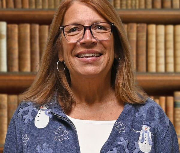 Annette Overmyer smiling in front of a library bookshelf, wearing glasses and a blue snowflake cardigan with holiday pins
