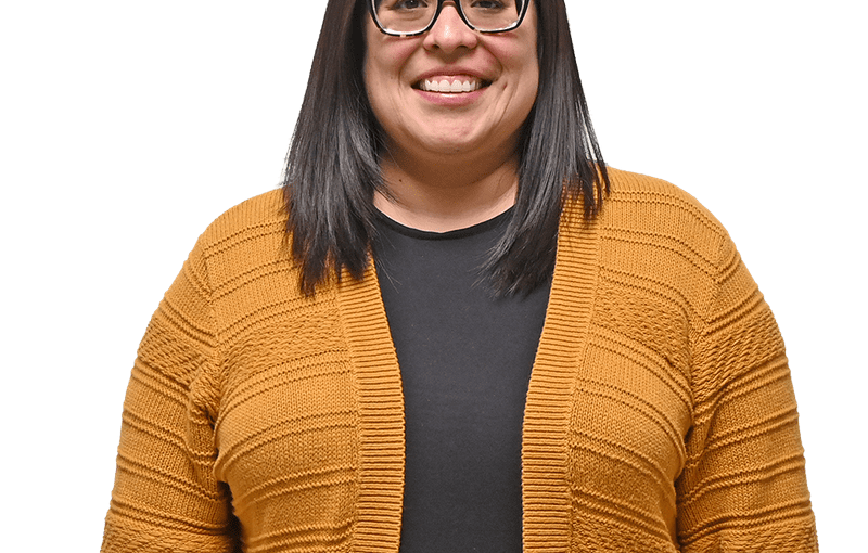 Clarissa McDermitt, Service and Support Administrator, smiling at camera wearing glasses and a mustard-colored cardigan over a black shirt