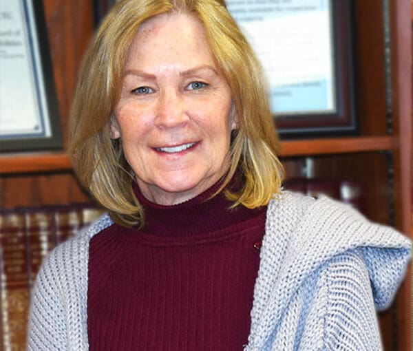 Woman with shoulder-length blonde hair wearing a burgundy turtleneck and light cardigan, smiling in an office setting with bookshelves and framed certificates in background