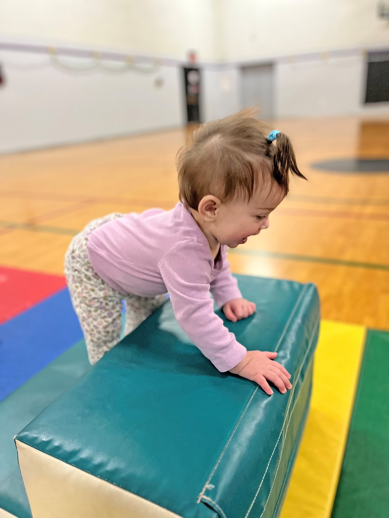 Toddler in pink shirt crawling on colorful foam mat during early intervention therapy session in gymnasium