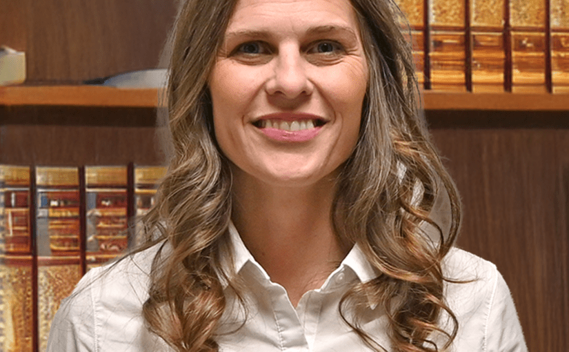 Professional woman with long wavy brown hair wearing a white button-up shirt, smiling in front of law books in an office setting