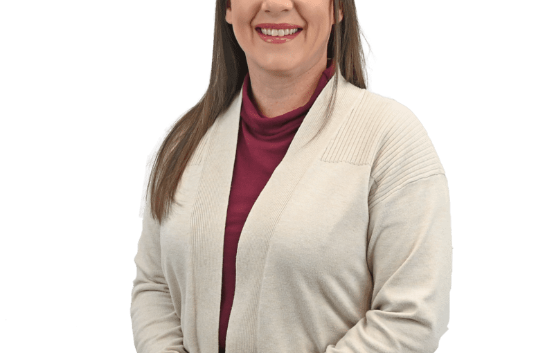 Kylie Paquin, Quality Assurance RN, smiling while sitting in a cream cardigan and maroon top against a white background
