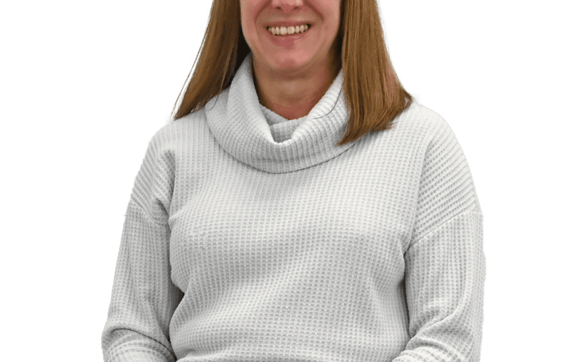 Lori Ohms, a smiling woman with reddish-brown hair wearing a light gray cowl neck sweater and blue jeans, sitting against a white background