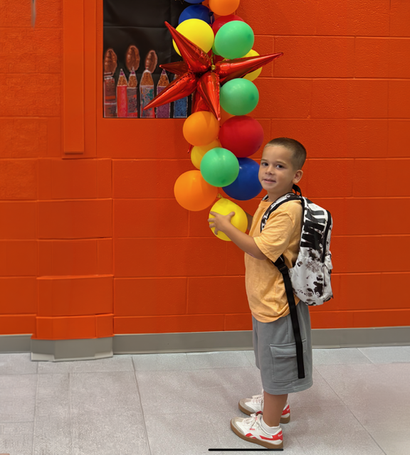 Young boy in yellow shirt and backpack smiling next to colorful balloon decoration on red brick wall