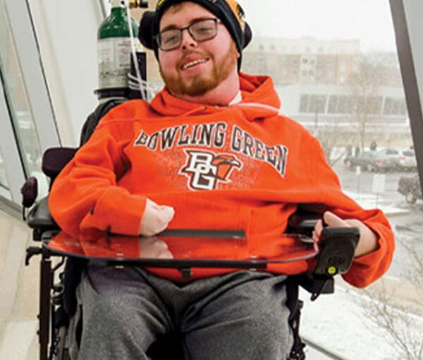 Man in wheelchair wearing orange Bowling Green hoodie and glasses, smiling at camera in front of large window overlooking city