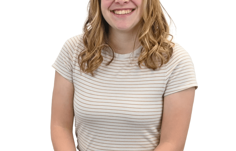 Smiling young woman with reddish-brown wavy hair and glasses wearing a striped beige and tan shirt and blue jeans