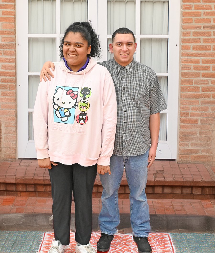 Two smiling teenagers standing together outside a brick building, one wearing a pink Hello Kitty hoodie and the other in a gray button-up shirt