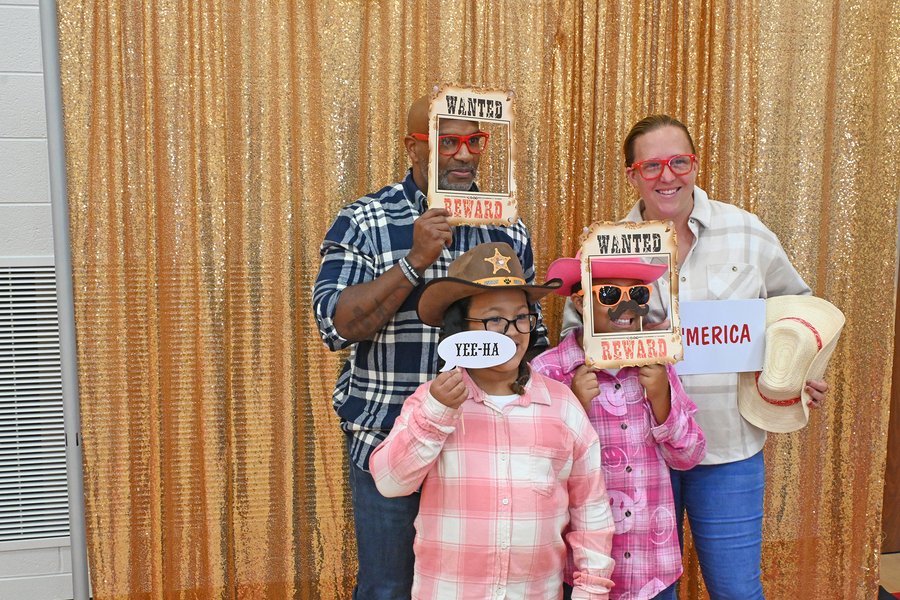 Four people wearing western-themed photo booth props including wanted posters, cowboy hats, and sunglasses in front of a gold sequin curtain