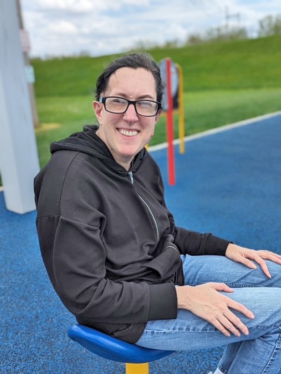 Smiling woman wearing glasses and black hoodie sitting on blue playground equipment with green field in background