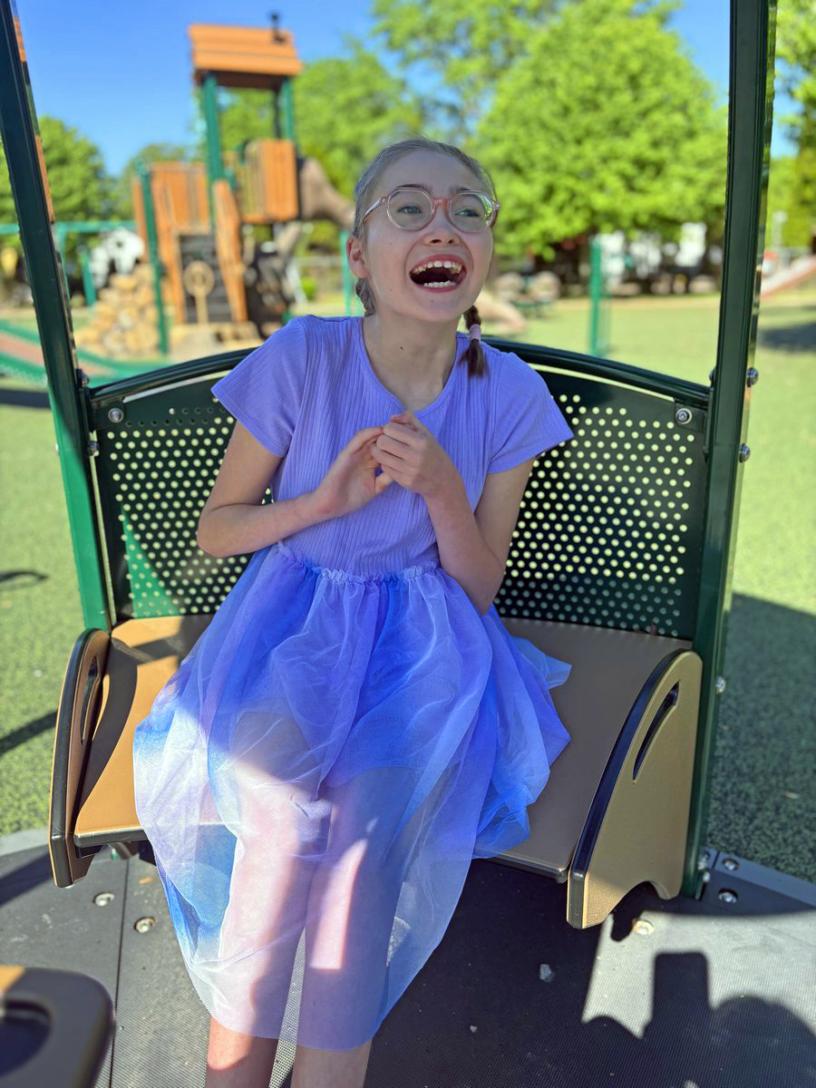 Girl with glasses smiling joyfully while sitting in a playground tube slide at a park with play equipment visible in background