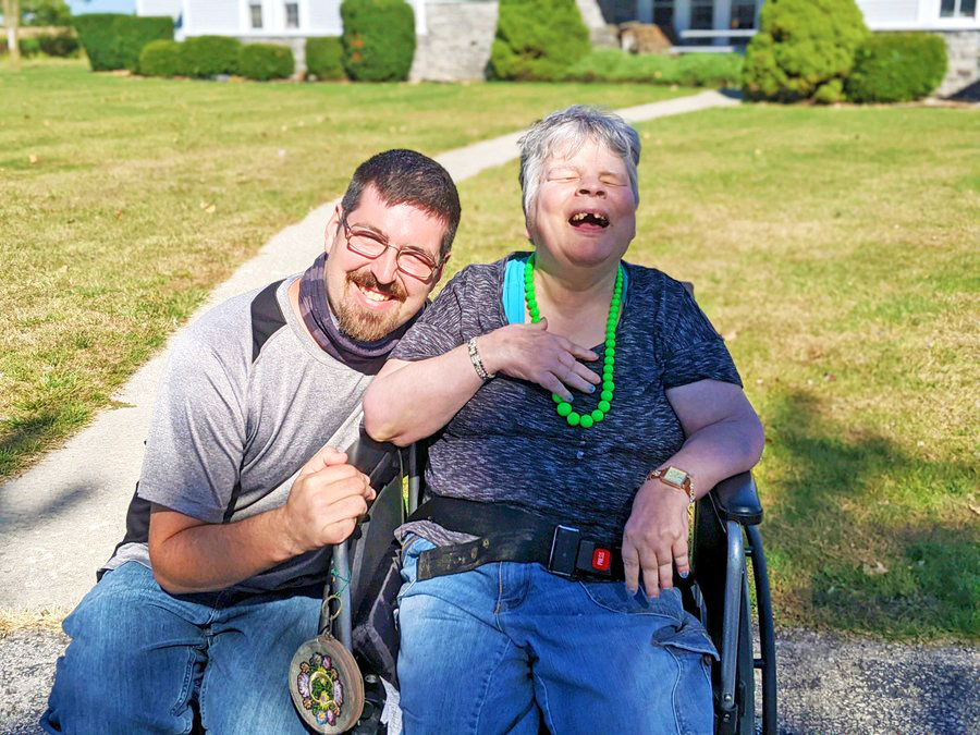 Two men smiling outdoors on a residential driveway in Sandusky County Ohio, one standing beside a wheelchair-using individual wearing a green beaded necklace, both appearing joyful