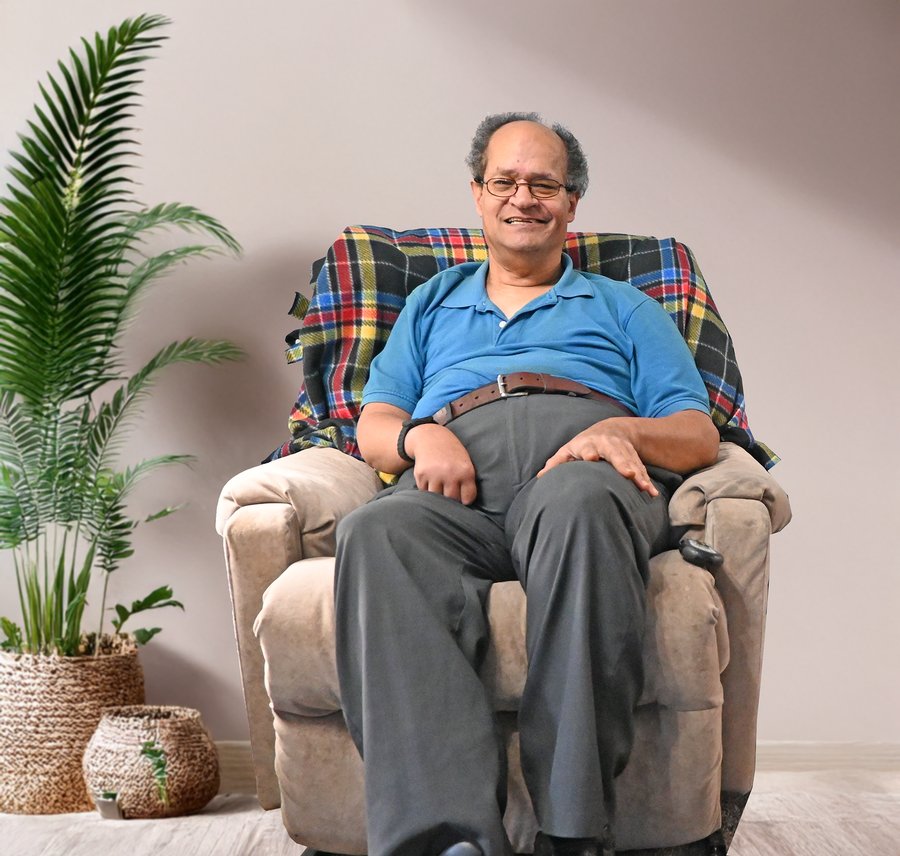 Middle-aged man with glasses wearing a blue polo shirt sitting in a beige armchair with plaid backing, potted plants beside him against a neutral wall