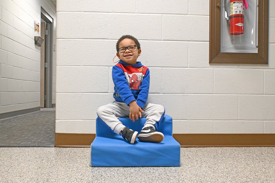 Young boy with glasses sitting on a blue foam block in a school hallway, smiling at the camera