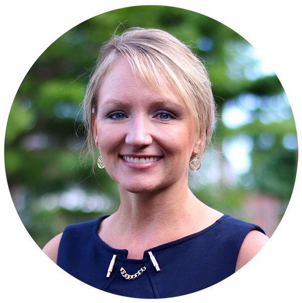 Professional headshot of a smiling woman with blonde hair wearing a dark blue sleeveless top with a chain necklace detail, photographed outdoors with a blurred green background