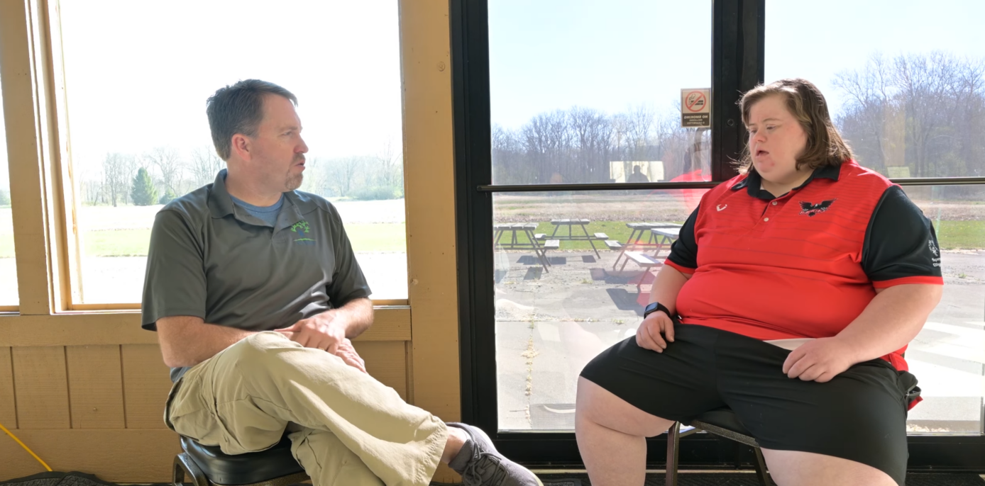 Two people having a conversation in an indoor setting with large windows overlooking a patio area and bare trees