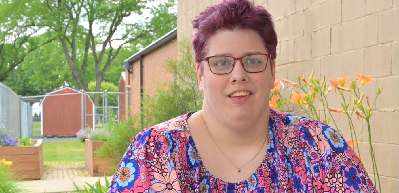 Person with purple hair and glasses smiling at camera in Sandusky County, Ohio garden setting with raised planters and brick building