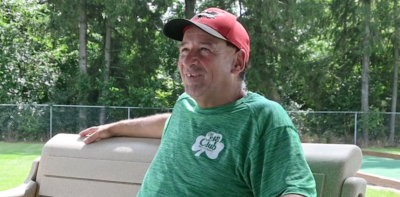 Man in green Big Club shirt and red cap smiling outdoors near mini golf course with trees in background
