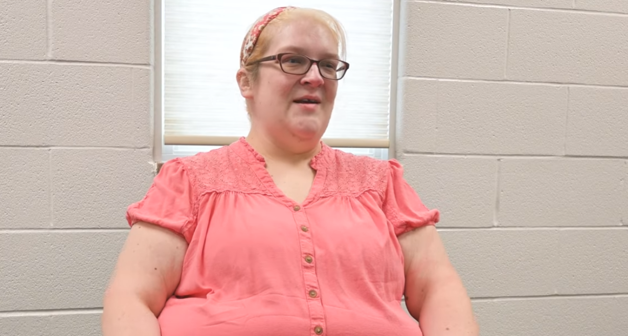 Woman wearing pink button-up shirt and glasses speaking indoors against brick wall background